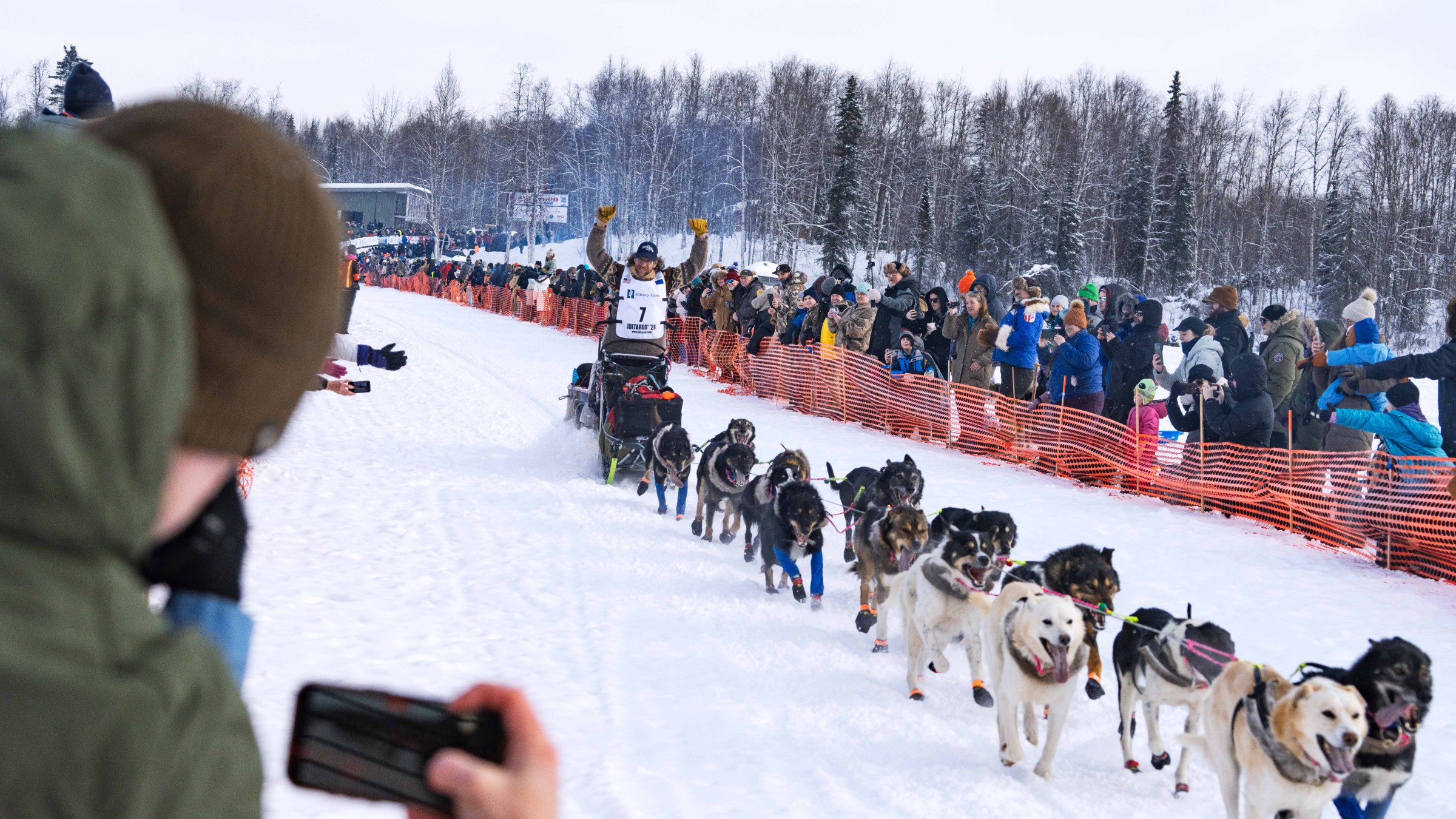 Dog sled race with crowd cheering, musher raising arms, snowy landscape.