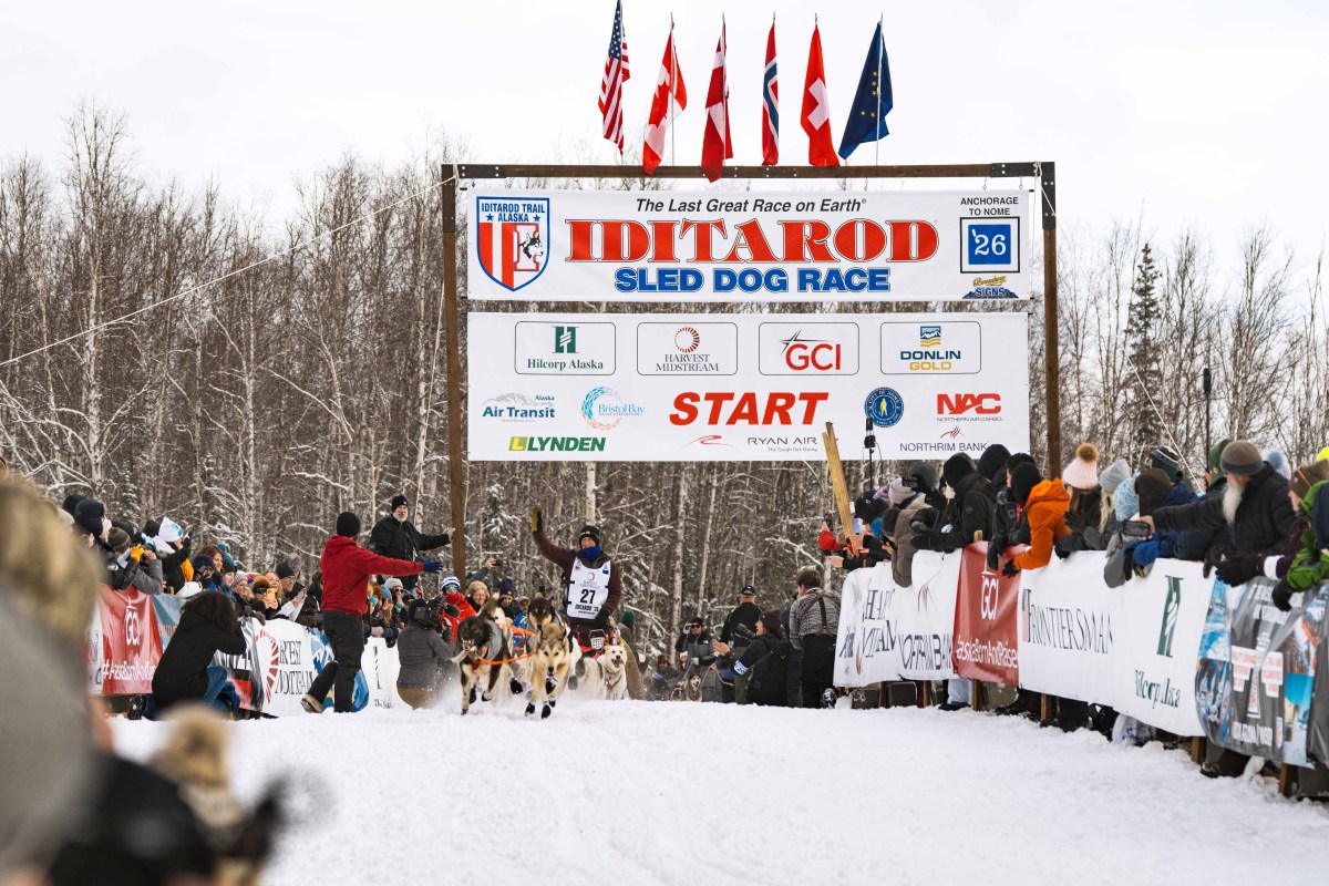 Musher and sled dogs start the Iditarod race surrounded by spectators and banners.