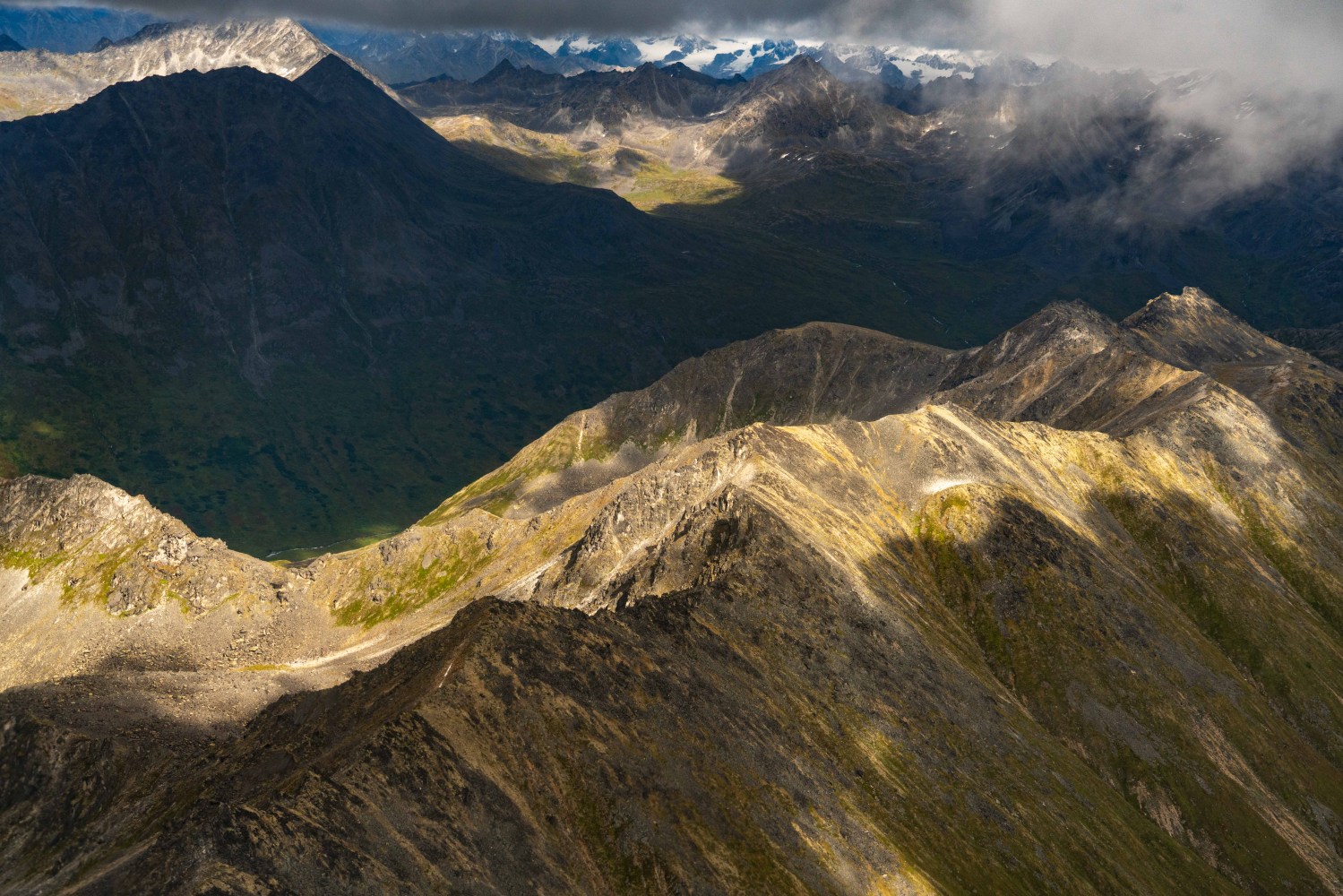 Aerial view of sunlit mountain range with shadows and cloudy sky.