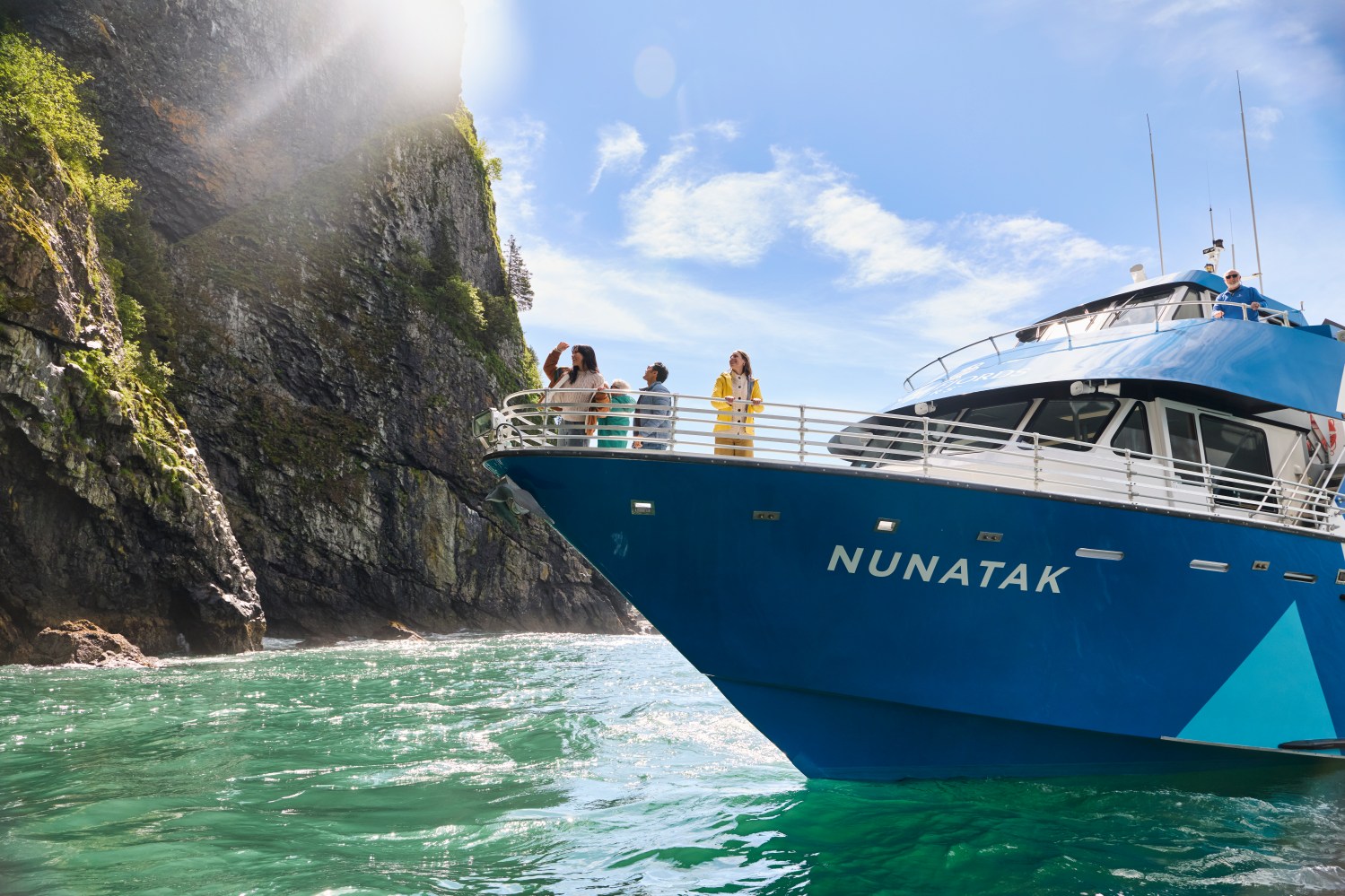 Tourists on a blue boat named NUNATAK near a rocky cliff under a bright sky.