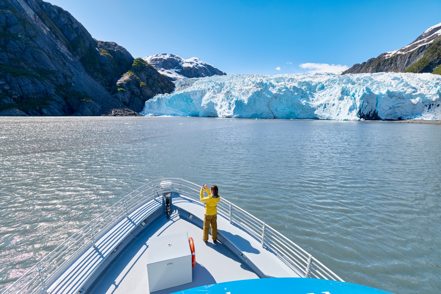 Boat approaches glacier with person in yellow jacket taking photo on deck, surrounded by mountains and blue sky.