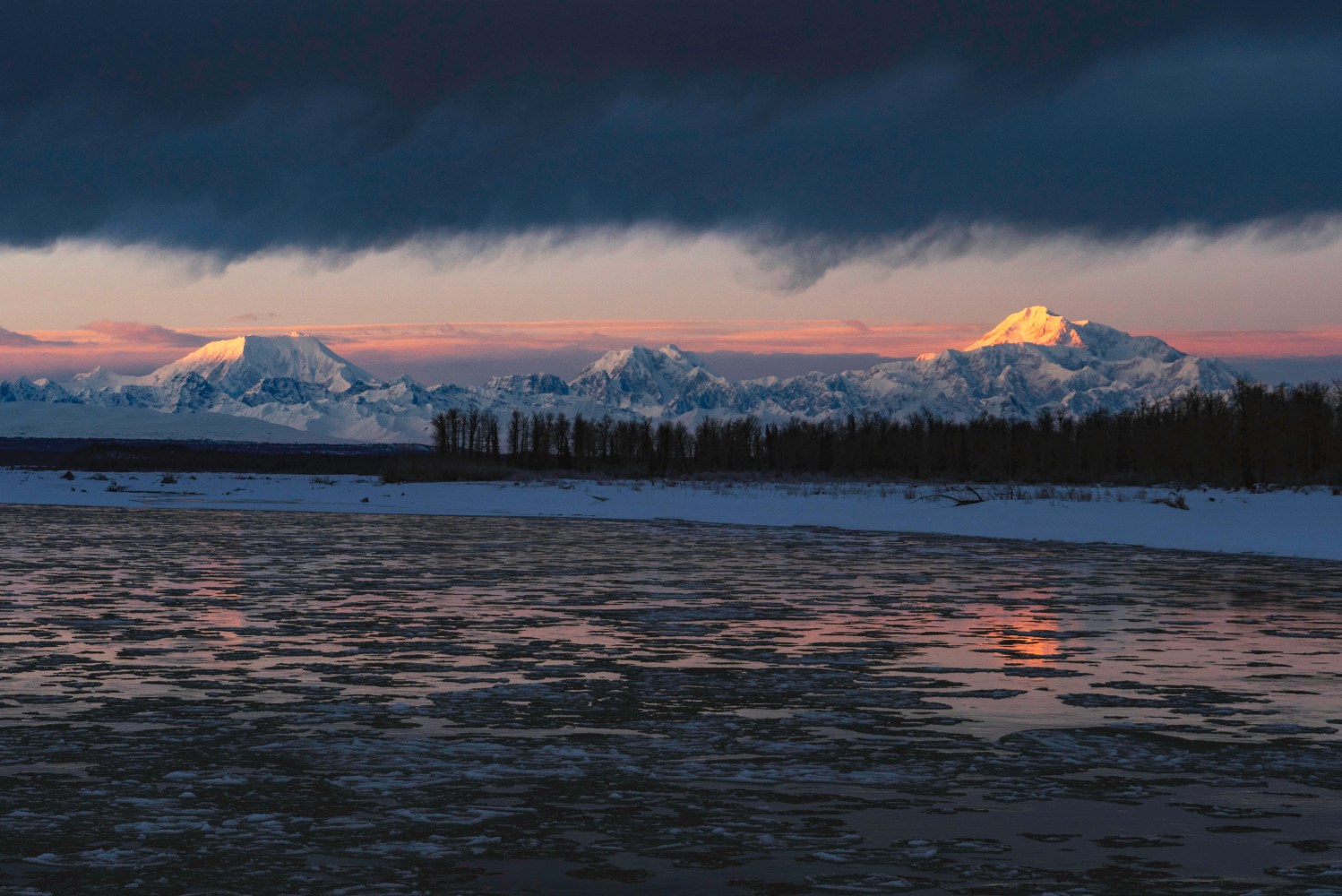 Snow-capped mountains at sunset with a river and dark clouds above.