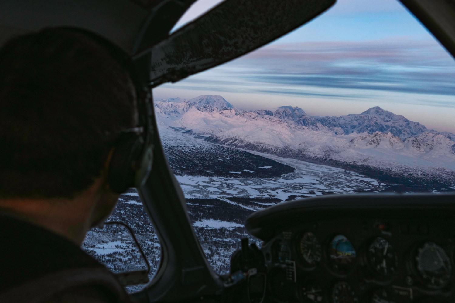 Pilot in cockpit flying over snowy mountains at sunrise featuring Denali and the Alaska Range