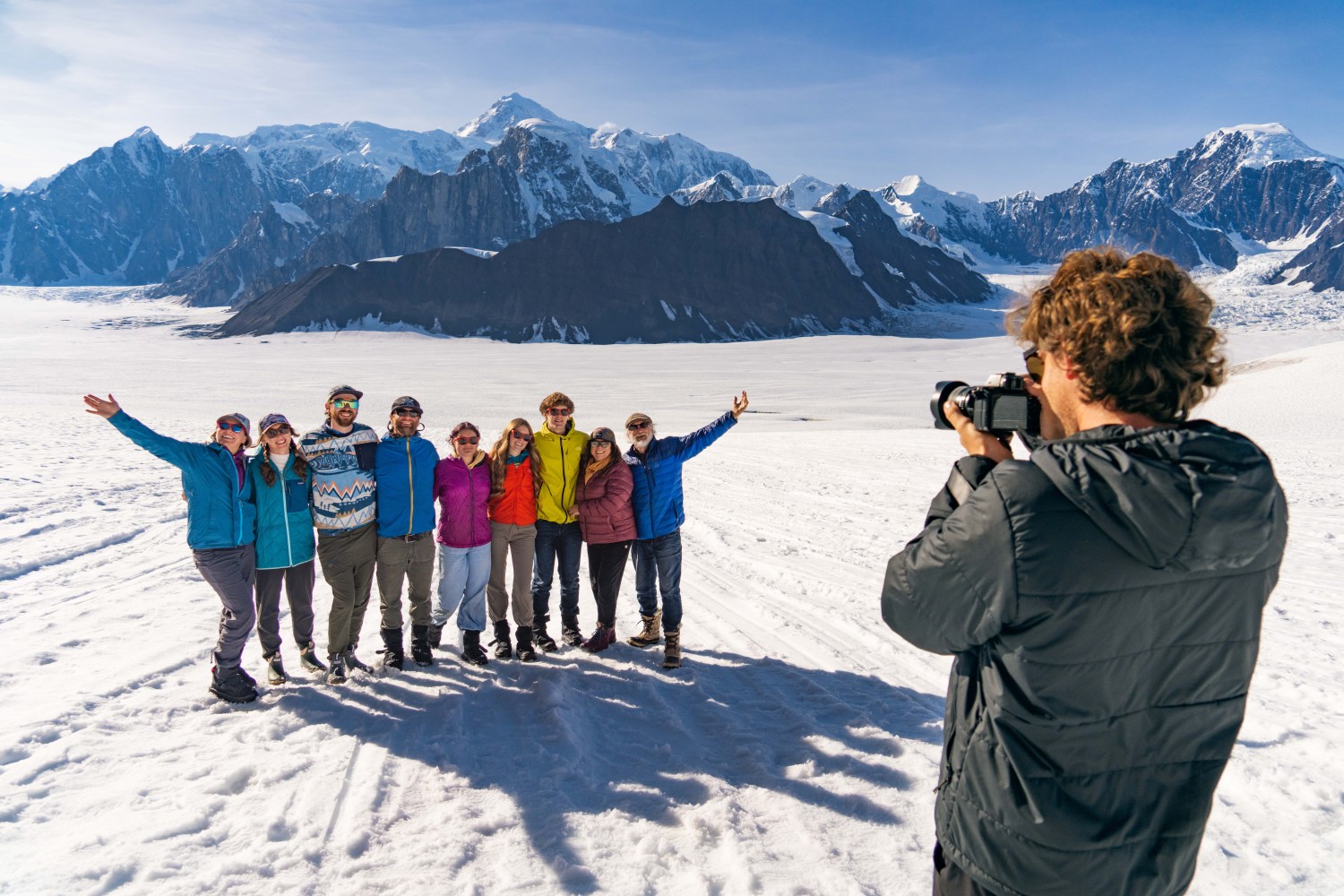 Group of people posing, a photographer captures them on a snowy landscape with mountains in the background.